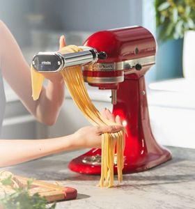 Una batidora KitchenAid color rojo cereza con el aditamento de Molino de Pasta preparando fideos.