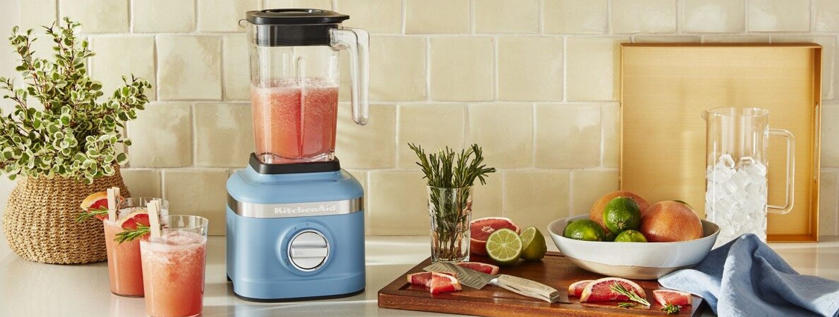 A blue countertop blender surrounded by fruit, cups of juice and an ice pitcher.