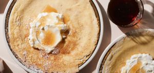 Plates of perfectly toasted crepes sit, dusted with powdered sugar. Each has a big dollop of melting butter in the center while maple syrup sits ready on the side.
