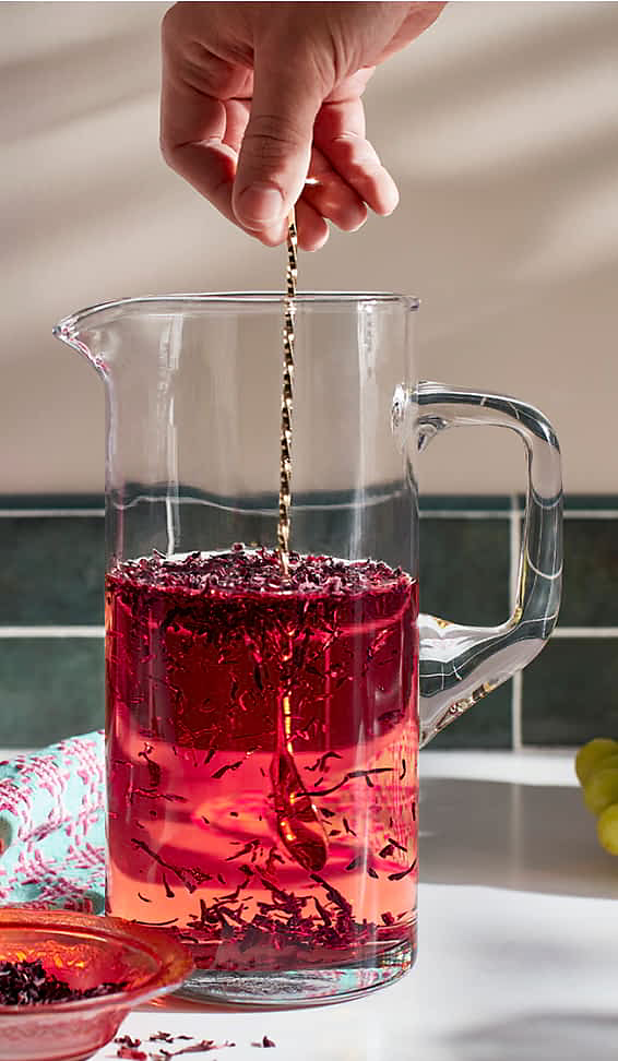 A pitcher filled with hibiscus leaves to make tea being stirred by a maker.
