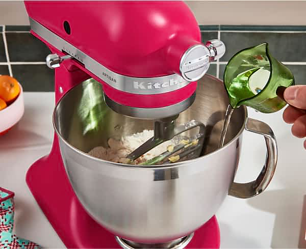 An Artisan® Series Stand Mixer in Hibiscus mixing dough, with a person pouring liquid into the mixer bowl.