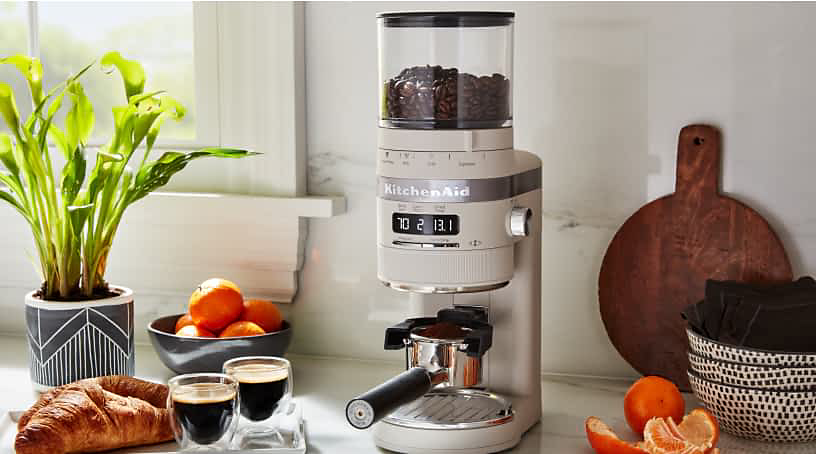 A KitchenAid® Burr Coffee Grinder in milkshake on a countertop, with espresso cups, fruit and pastries around the appliance.
