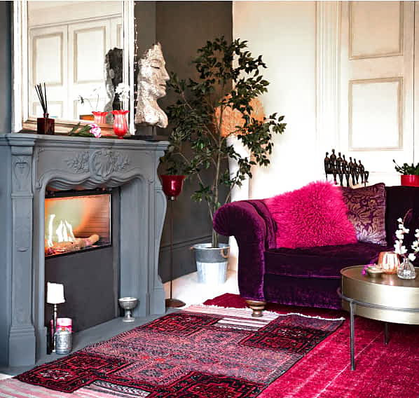 A living room with a lit fire in the fireplace and fuchsia accents across the couches, rugs and pillows.