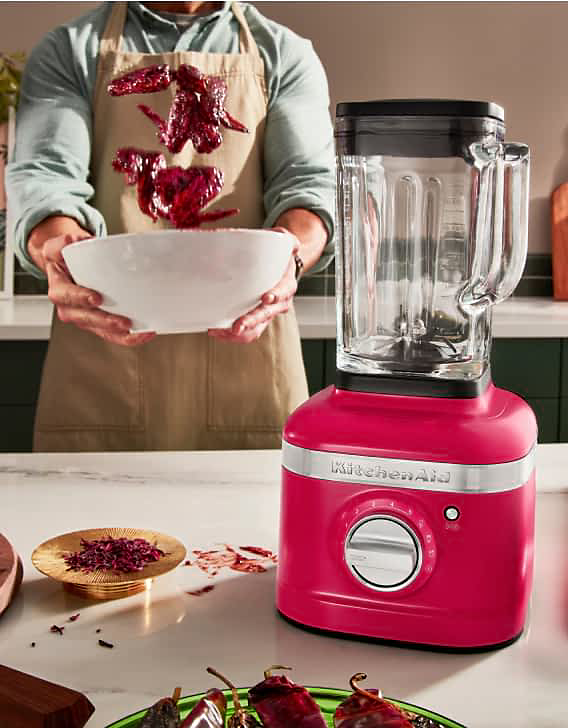 A person tossing hibiscus-coated chicken wings in a bowl with a K400 blender on the countertop.