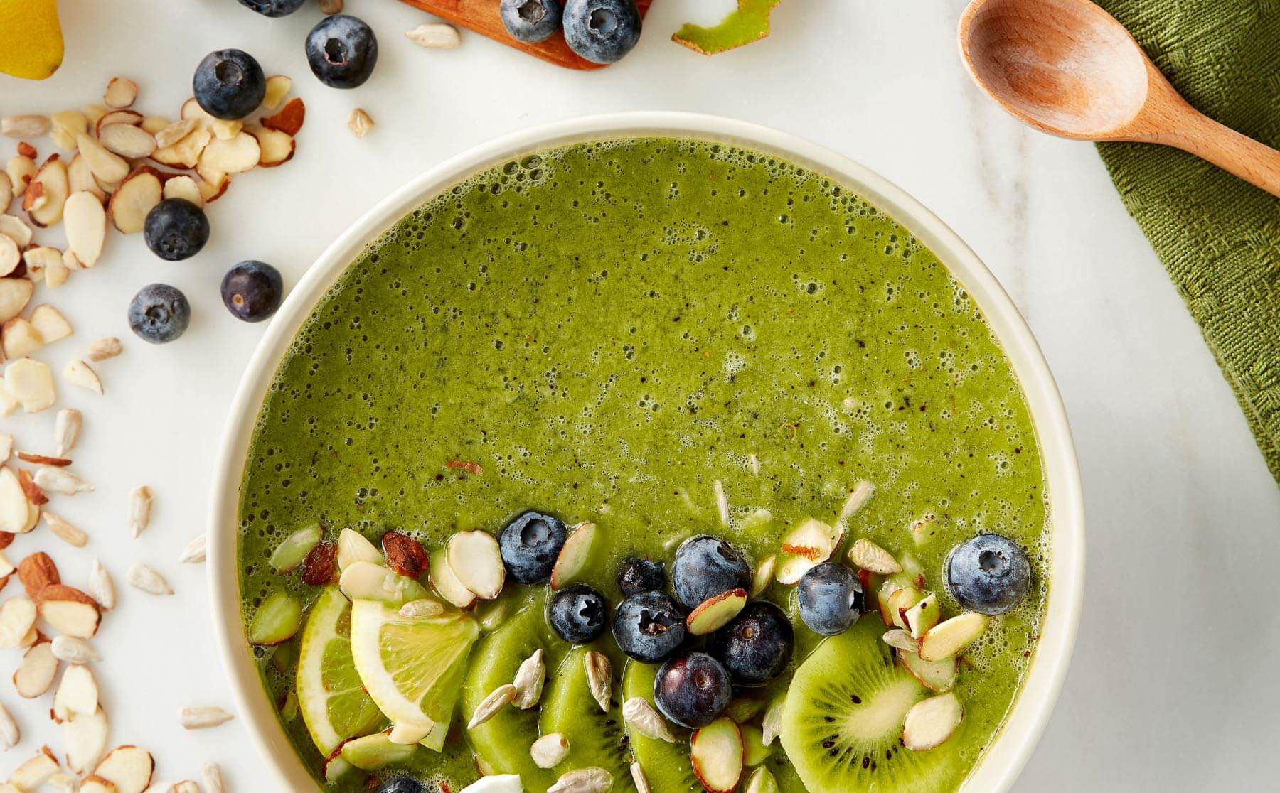 A white bowl of green smoothie topped with sliced kiwis, lemons, almonds and blueberries.