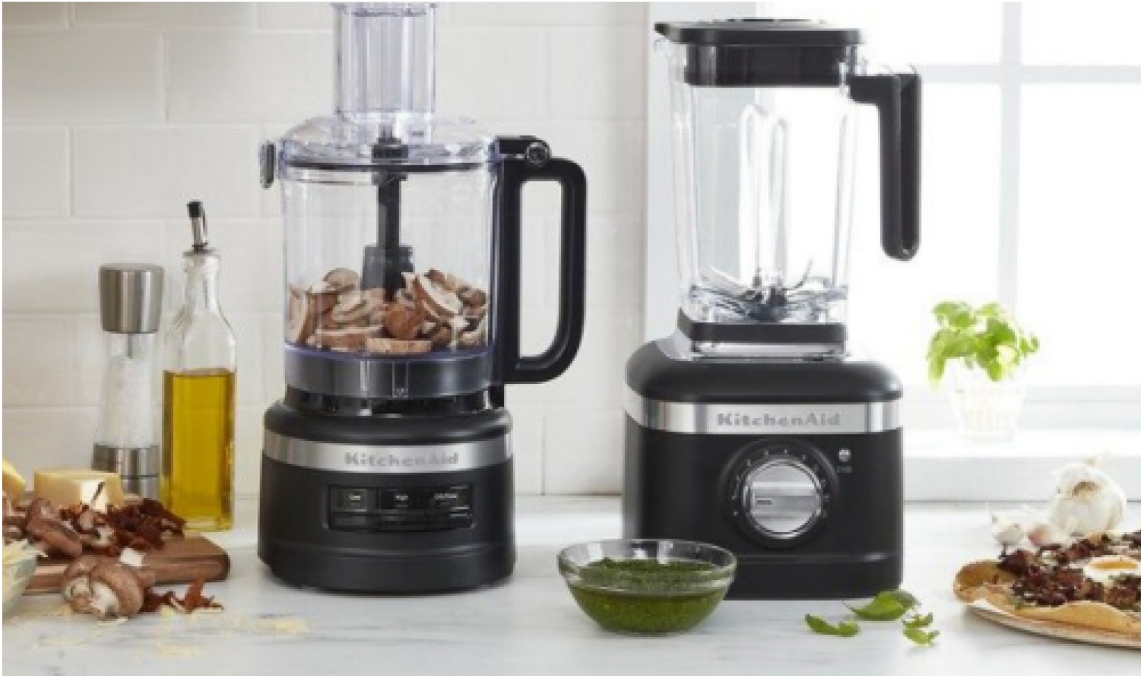 A KitchenAid food processor and blender on a countertop with different ingredients scattered around the counter. 