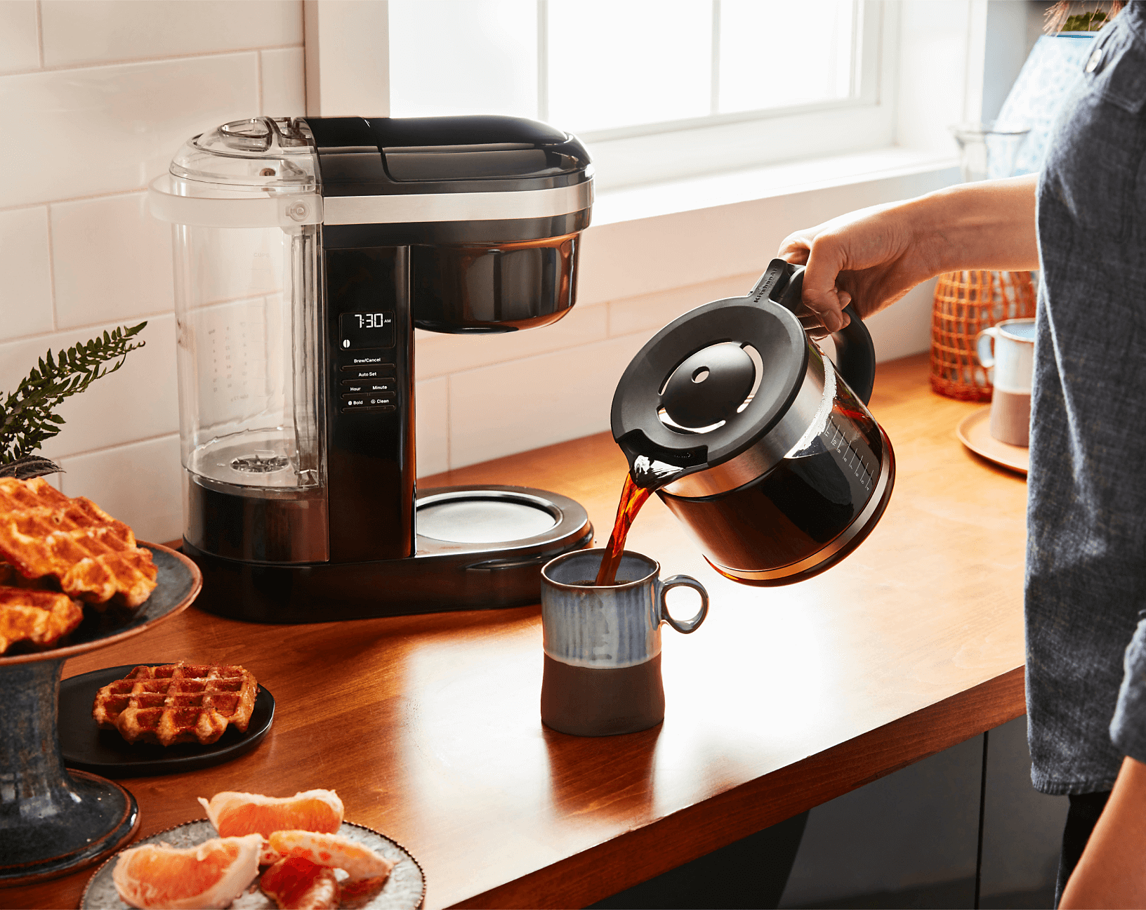 A person pouring coffee into a ceramic coffee mug on a countertop near a KitchenAid® drip coffee machine and different breakfast ingredients. 