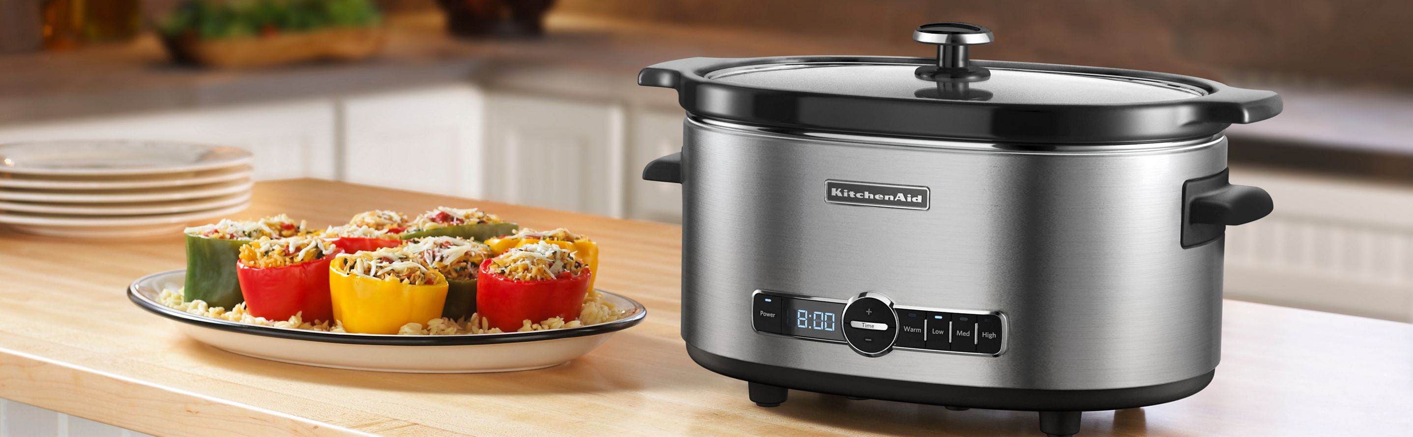 A KitchenAid slow cooker sitting on a countertop next to a plate of prepared stuffed peppers.