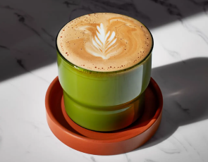A flat white served with latte art in a green glass on a burnt orange coaster.