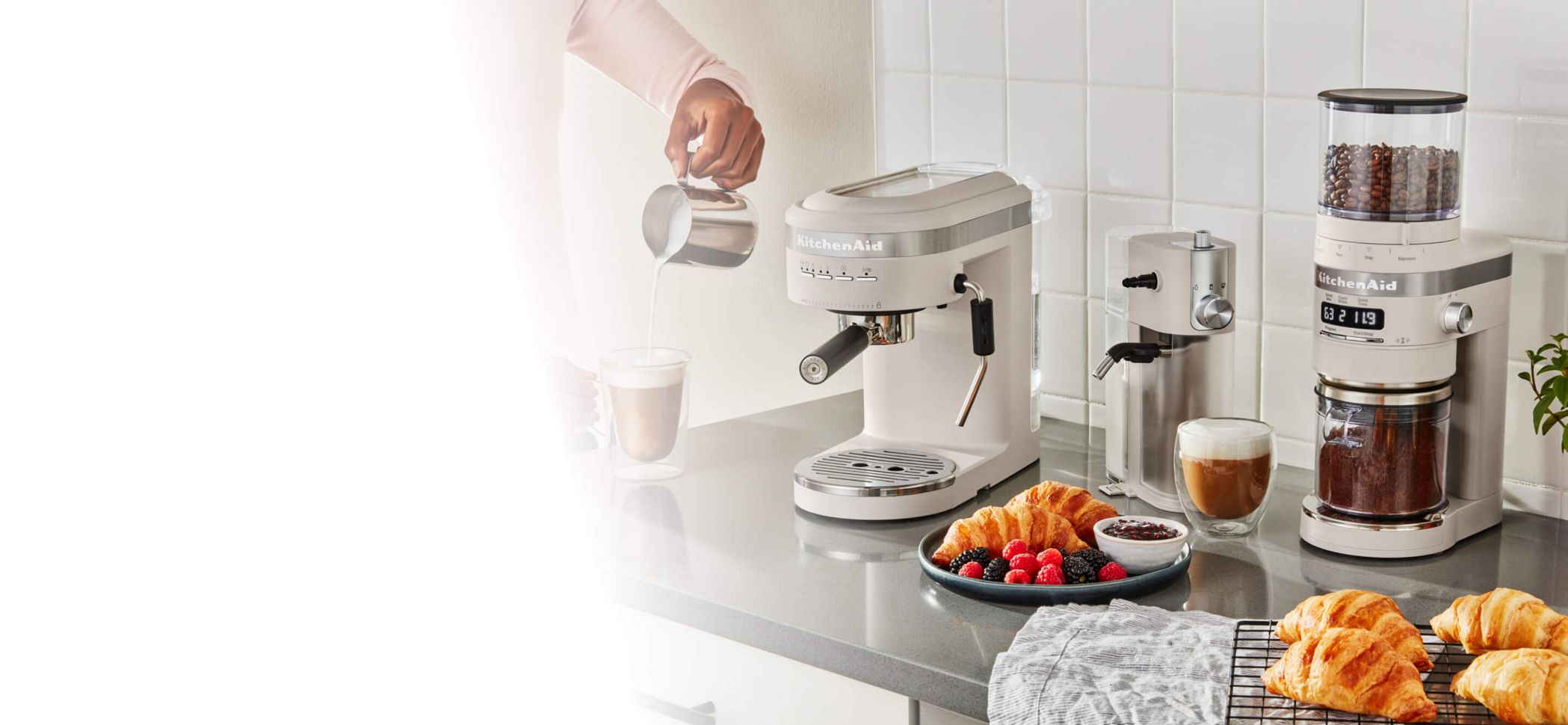 A KitchenAid® Espresso Machine, Milk Frother and Burr Grinder on a counter top with fresh espresso, fruits and croissants.