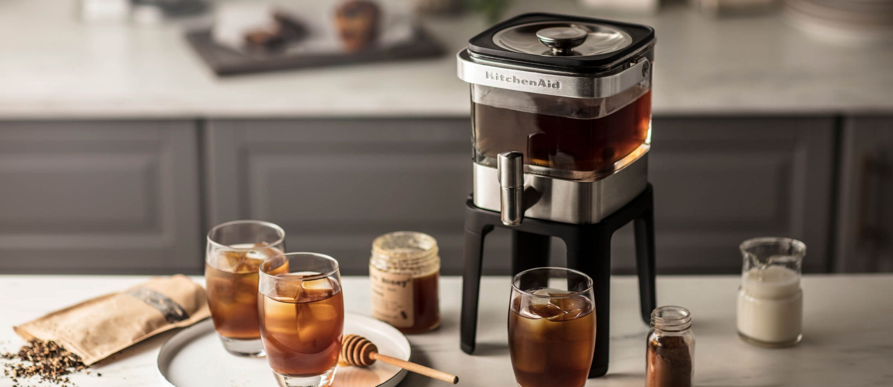 A KitchenAid® Cold Brew Maker with different cups and topping scattered on the countertop