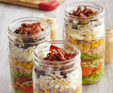 Three tall, glass mason jars filled with chopped ingredients for to-go salad.