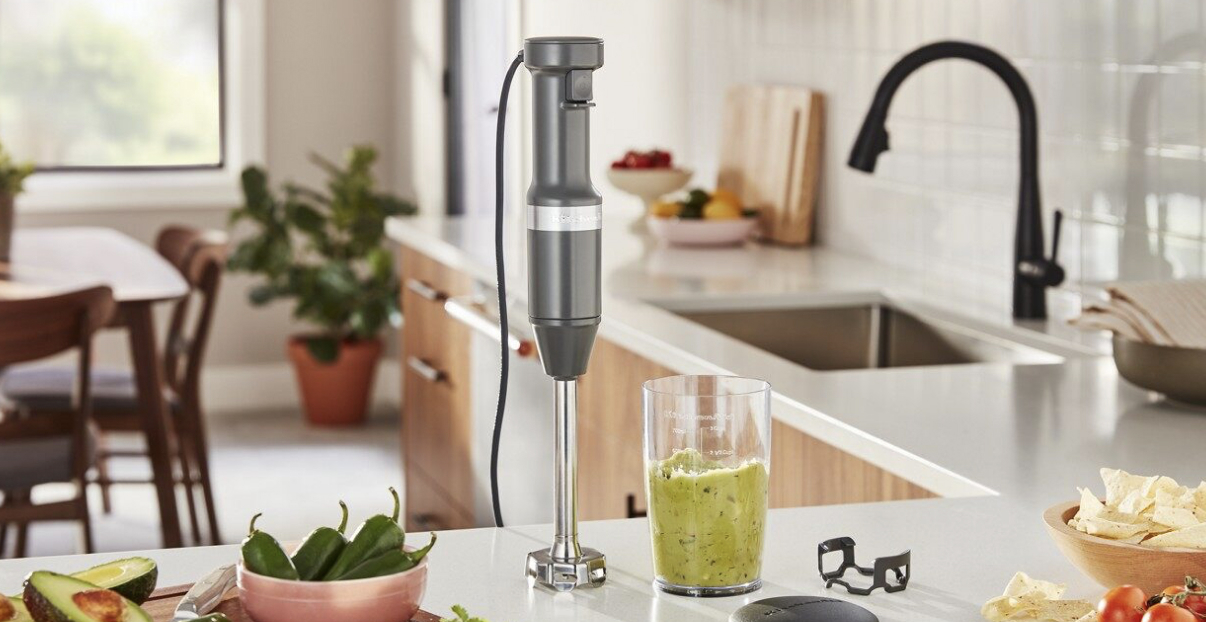 A hand blender with avocados and peppers on the counter.