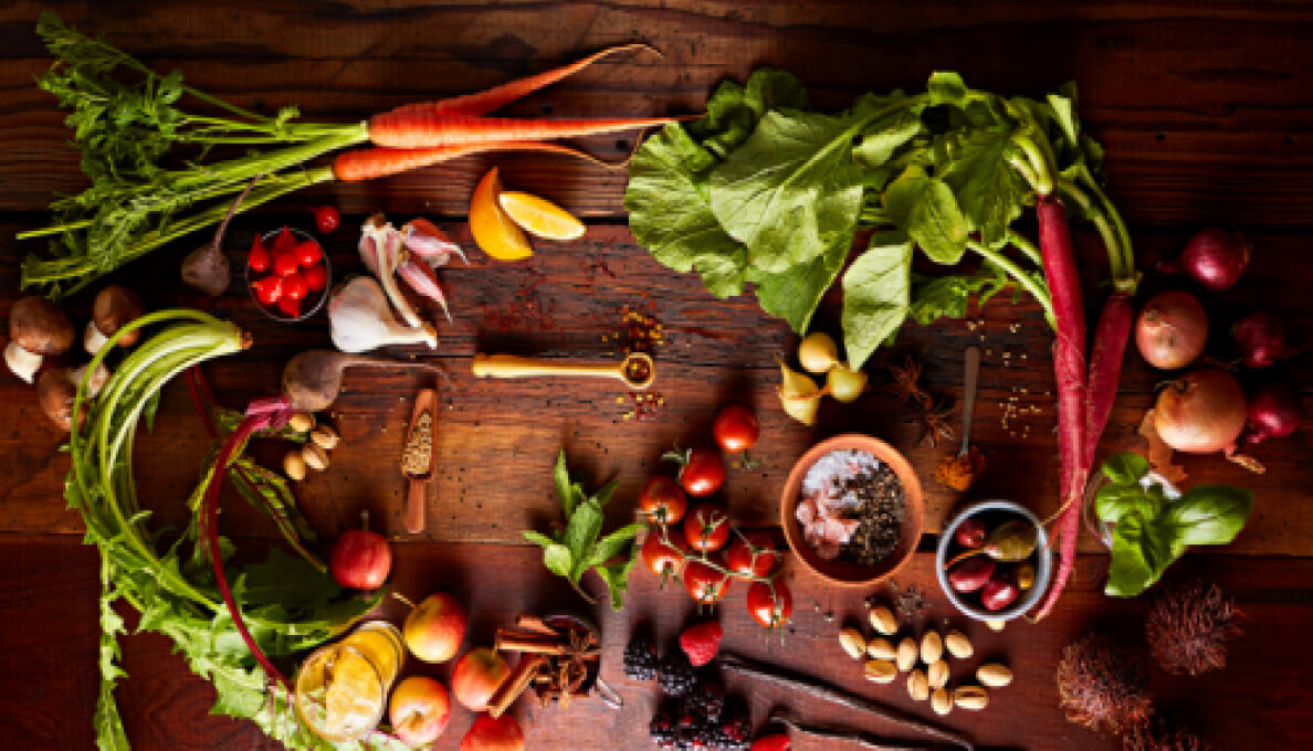 Whole spices, vegetables and fruits arranged on a wood countertop.