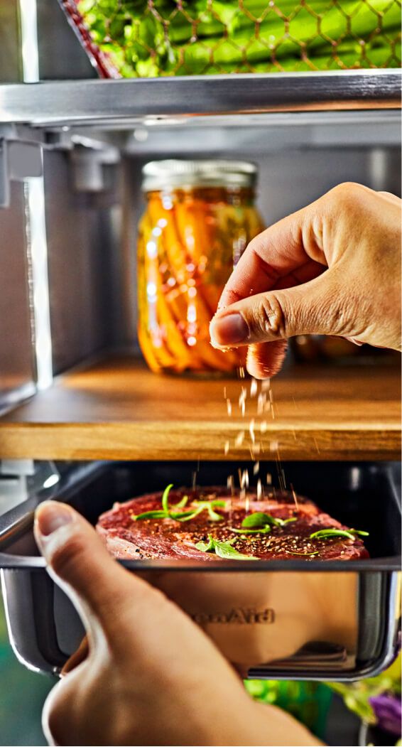 A person seasoning a steak straight from the refrigerator with basil and salt.