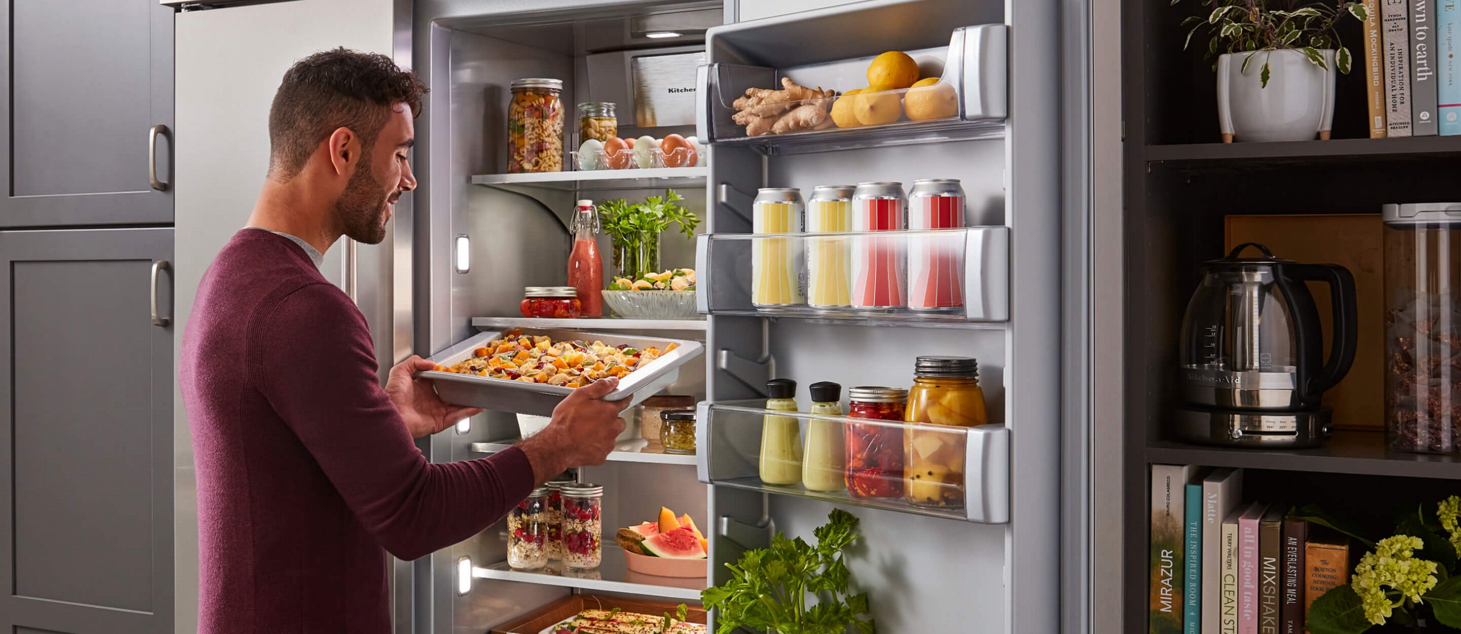 A person placing a sliding storage tray into a fully stocked KitchenAid® built-in refrigerator.