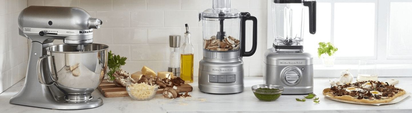 A KitchenAid® Stand Mixer, Food Processor and Blender on a white countertop, surrounded by different ingredients on a cutting board and a prepared breakfast pizza.