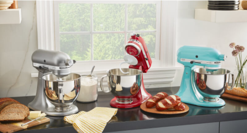 A trio of KitchenAid® Stand Mixers on a countertop with fresh baked bread on plates. 