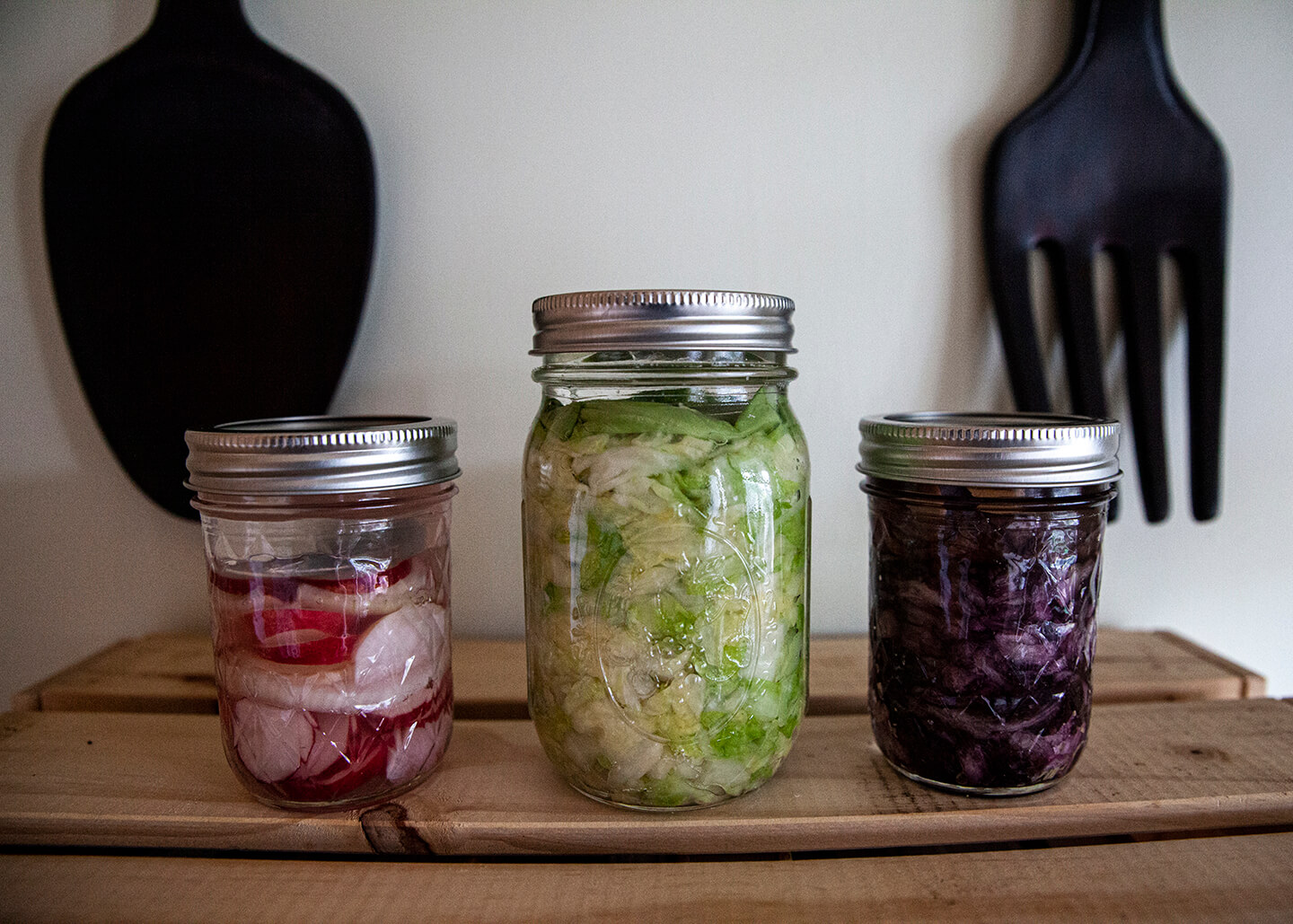Three glass jars holding fermented radishes, red cabbage and green cabbage.