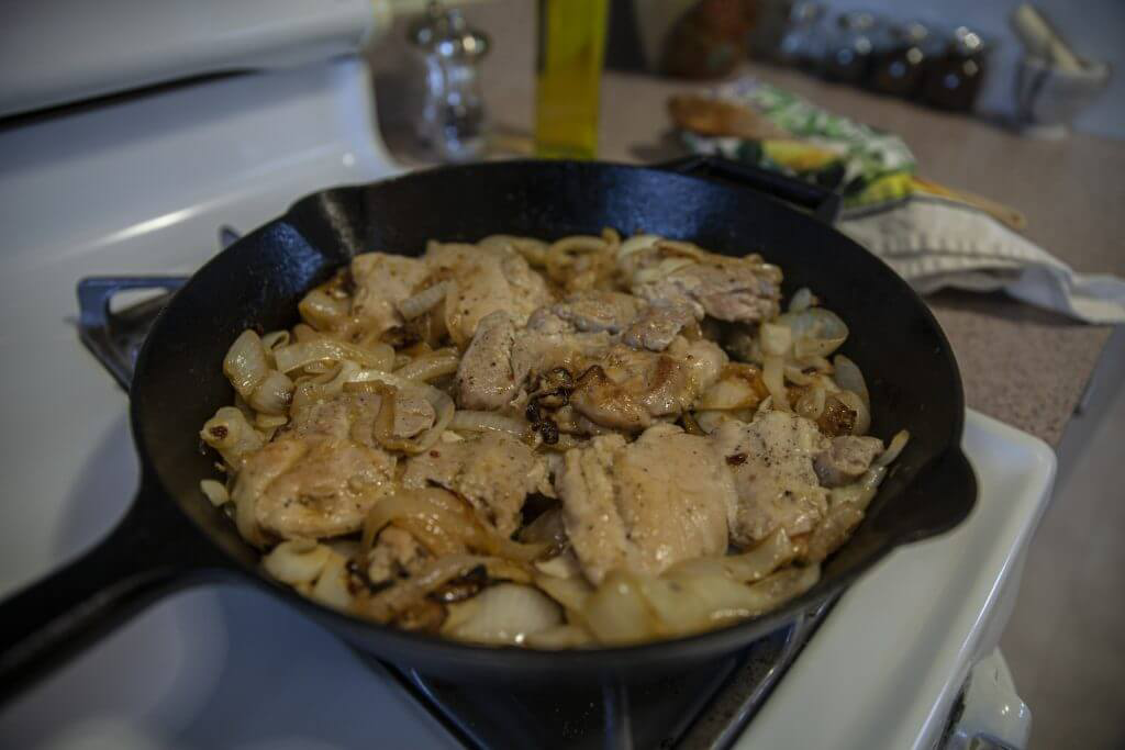 A stove with a cast iron skillet holding chicken thighs and caramelized onions.