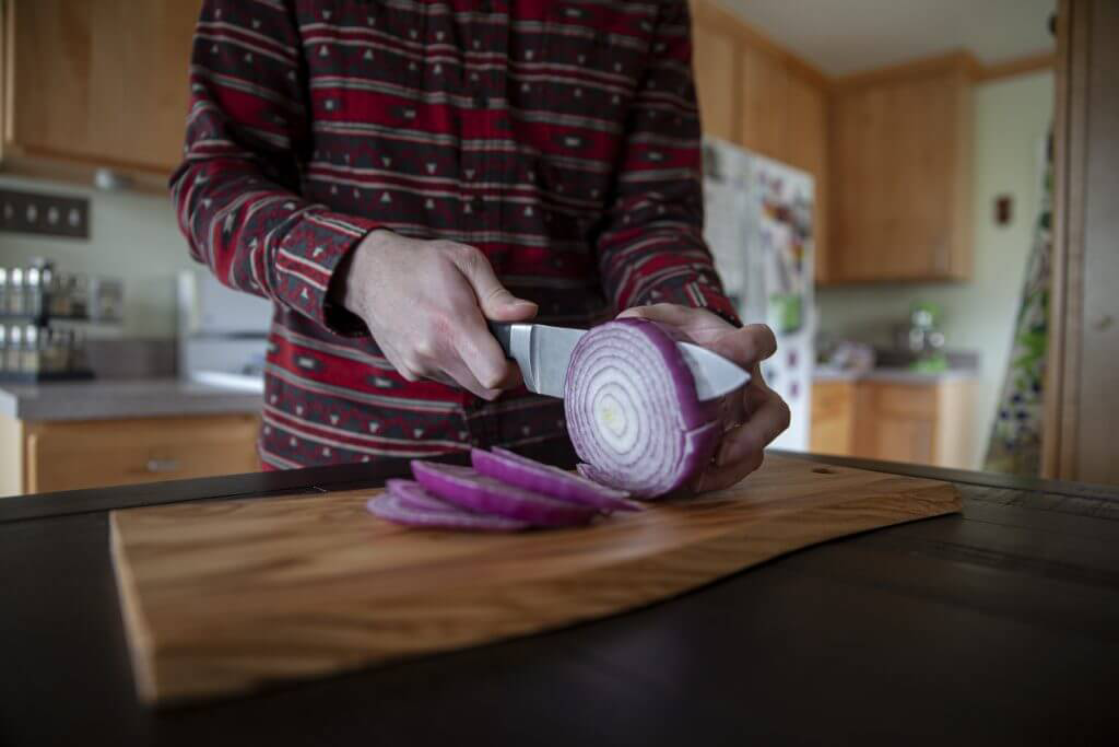 A person slicing a red onion on a wooden cutting board with a large knife.