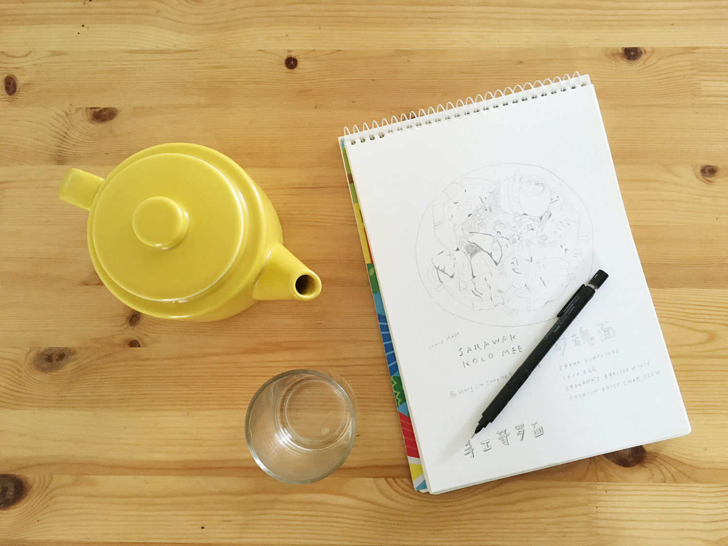 A sketch book resting on a table with a pot of tea.