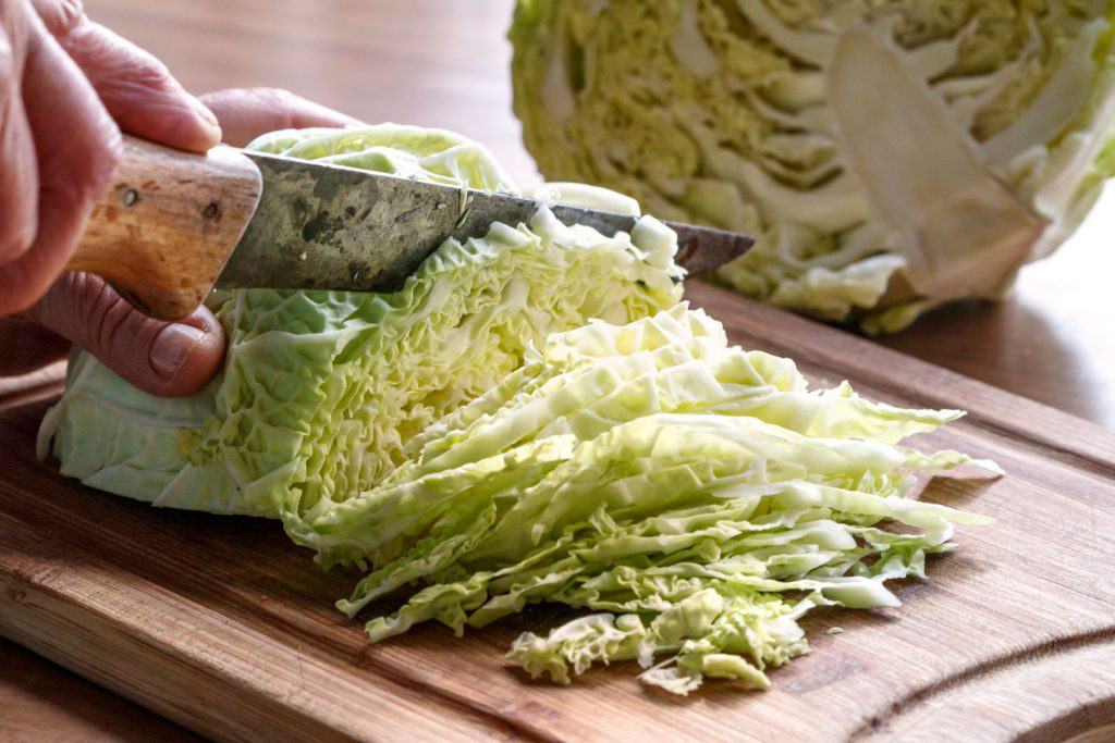 A person thinly chopping a halved cabbage on a wooden cutting board.