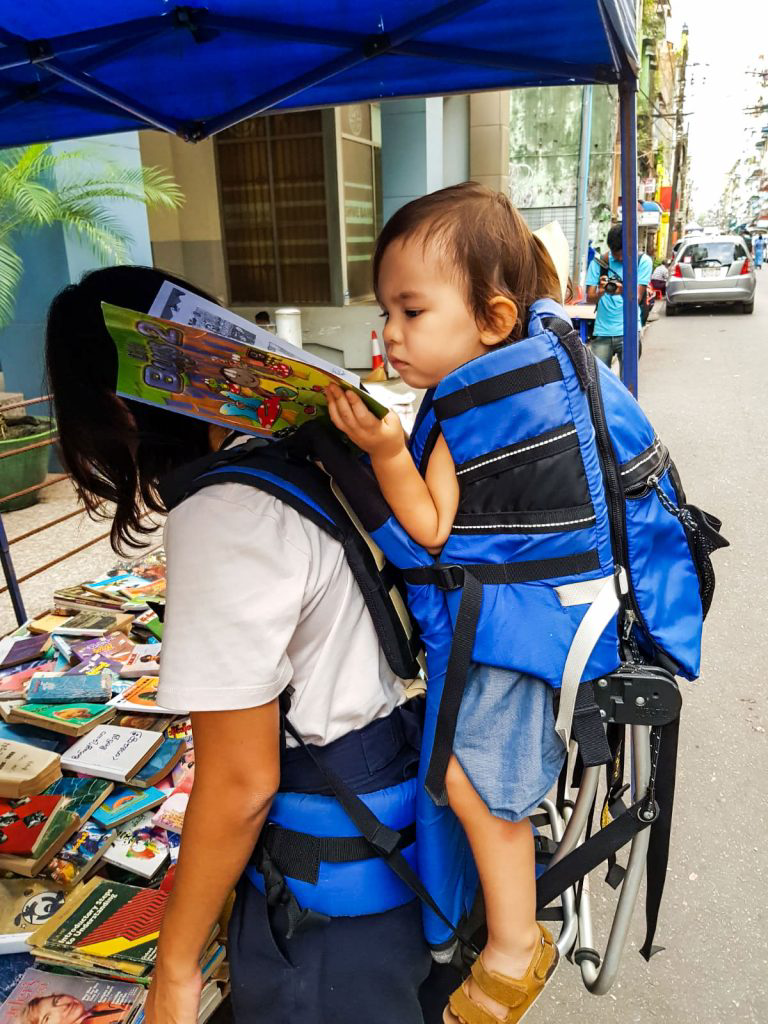 Wee Ling Soh's daughter reading a magazine while being carried in a blue backpack carrier.