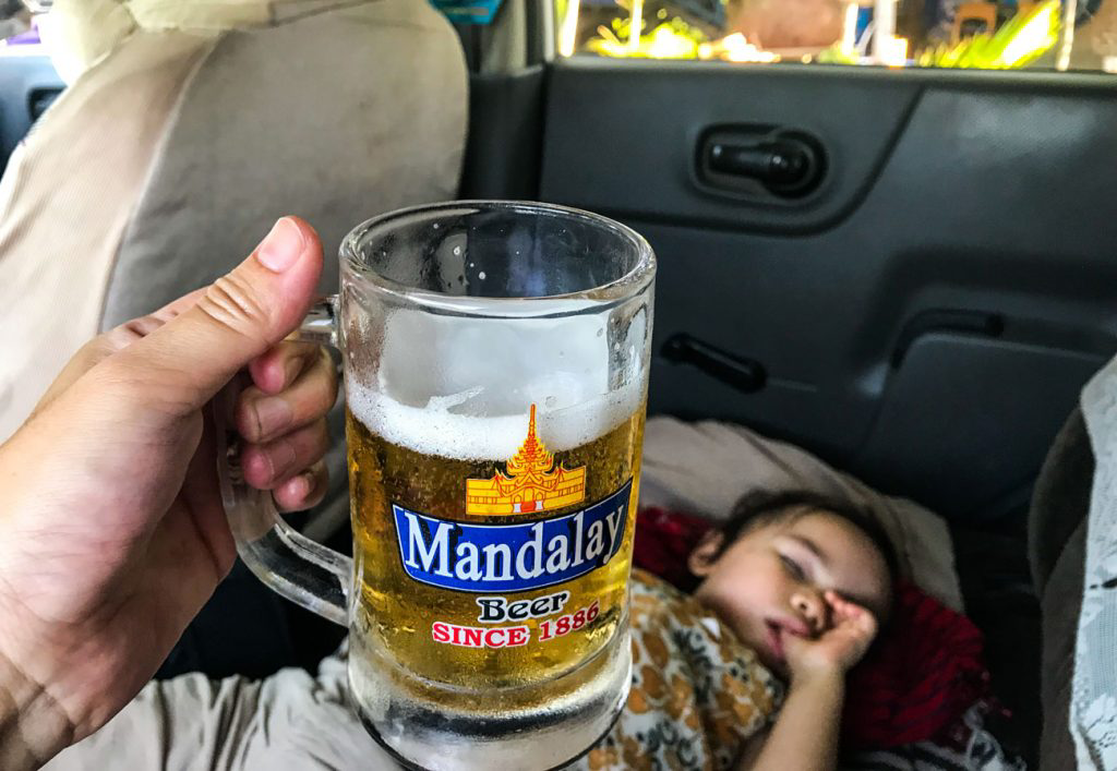 A hand holding a local lager in the back of a car with a sleeping child.