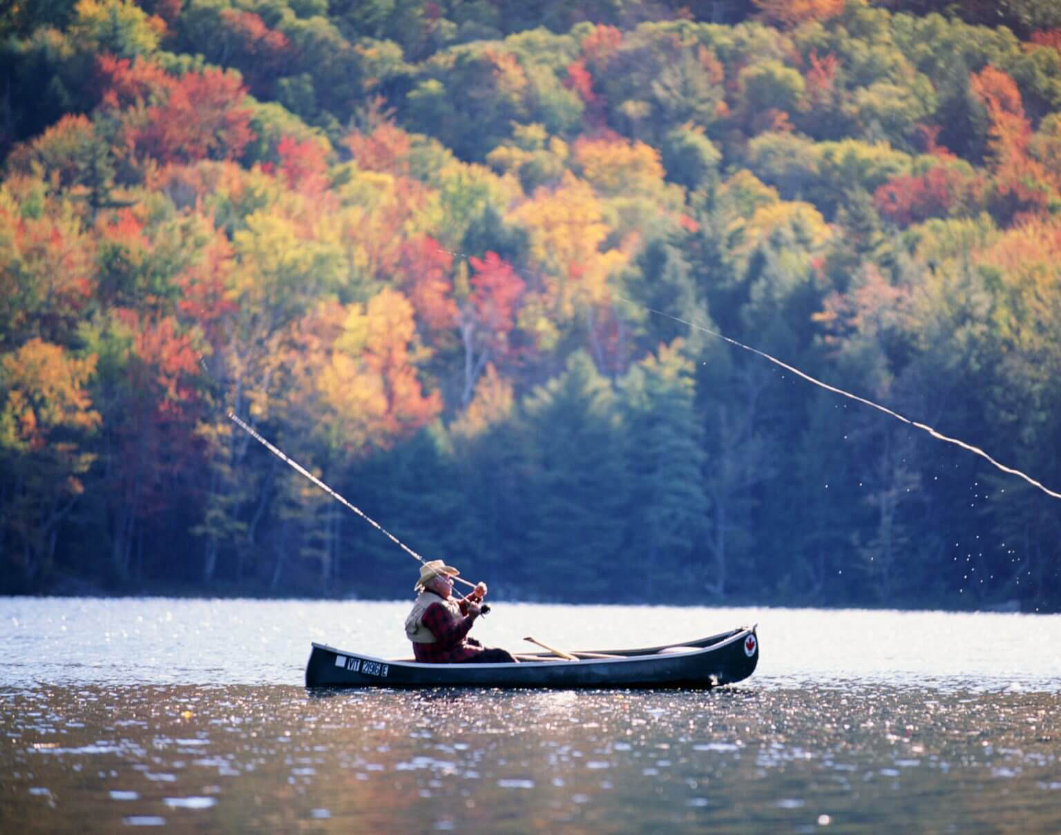 An elderly man in straw hat fishing in a canoe on a calm lake.