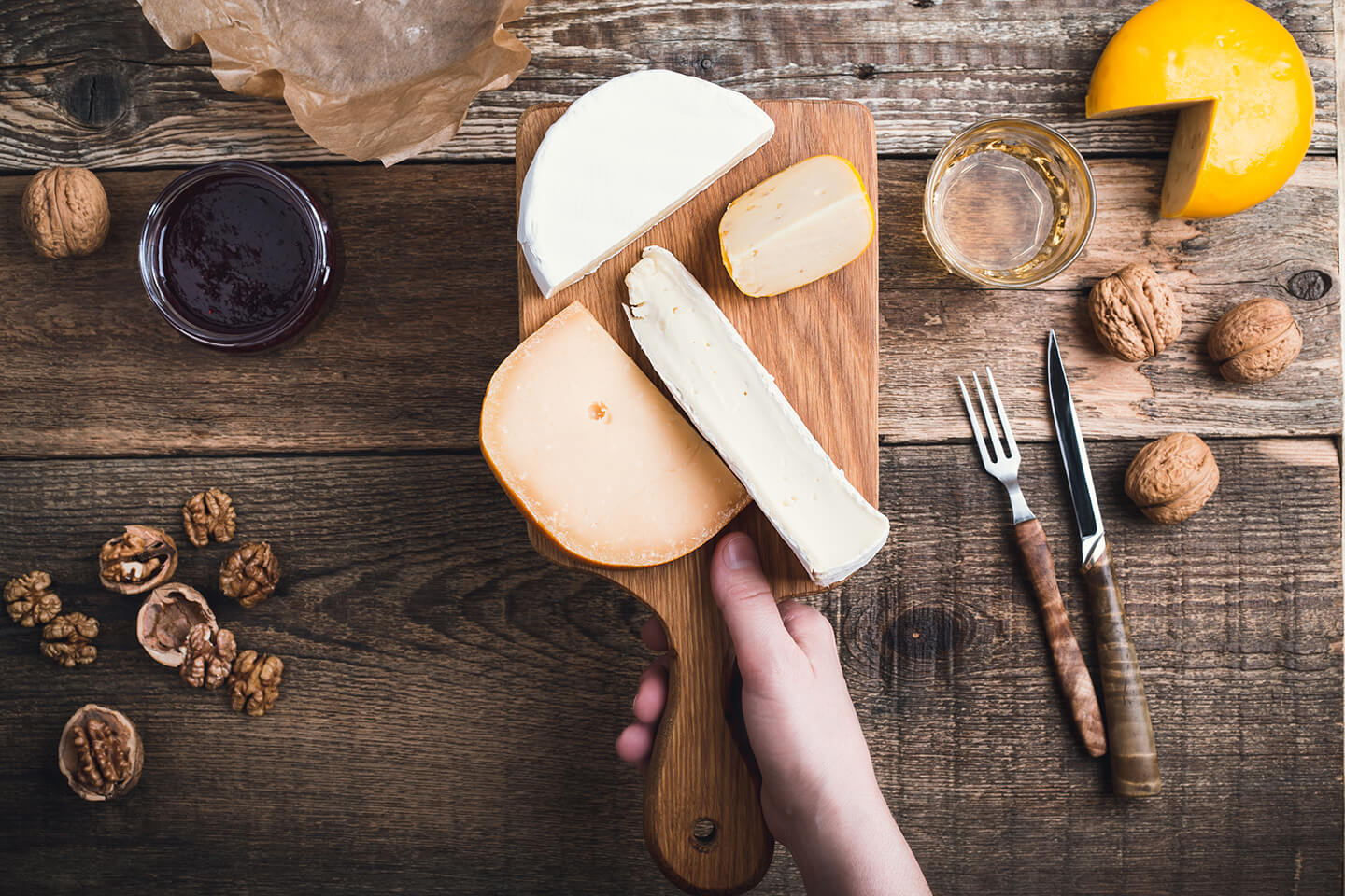 A hand holding up a rustic, wooden cheese board with assorted cheeses.