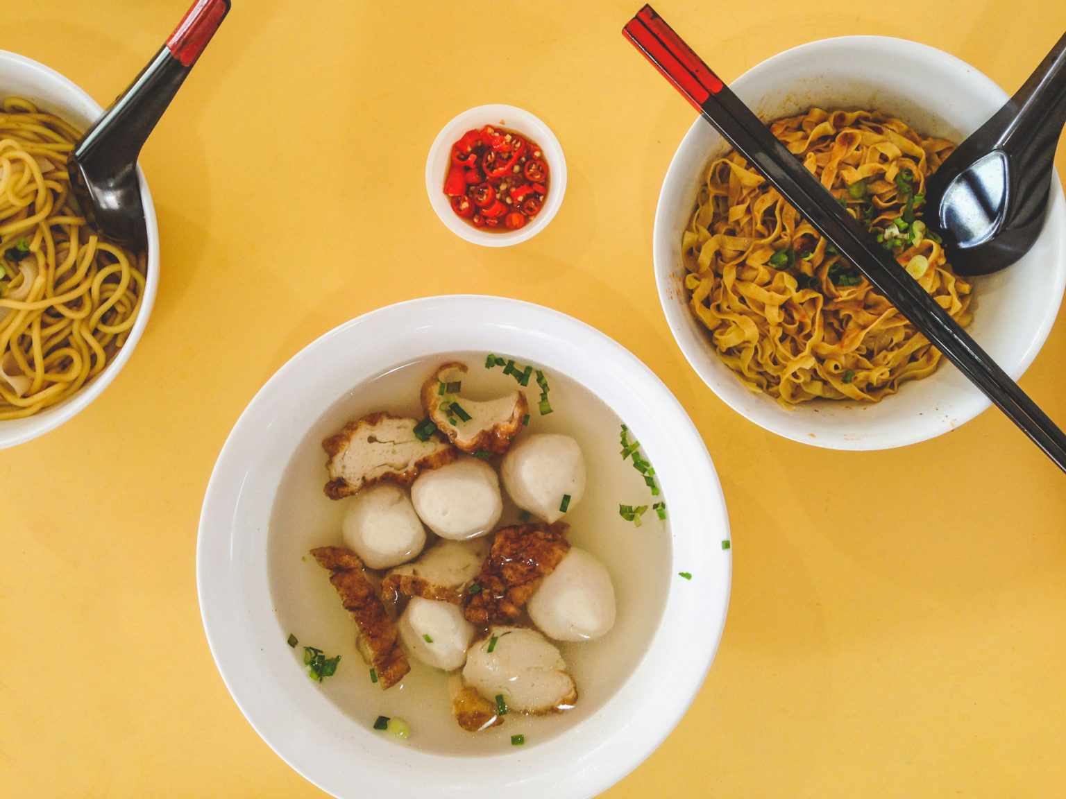 Bowls of Singaporean noodles and soup on a bright yellow table.