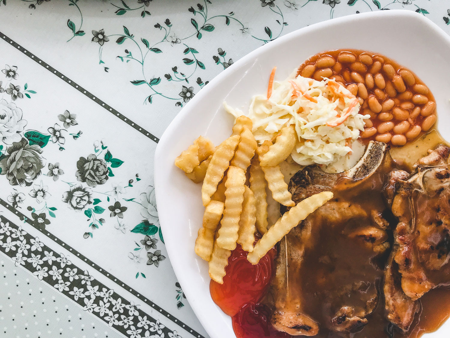 A plated porkchop in brown sauce served with crinkle-cut fries, baked beans and coleslaw.