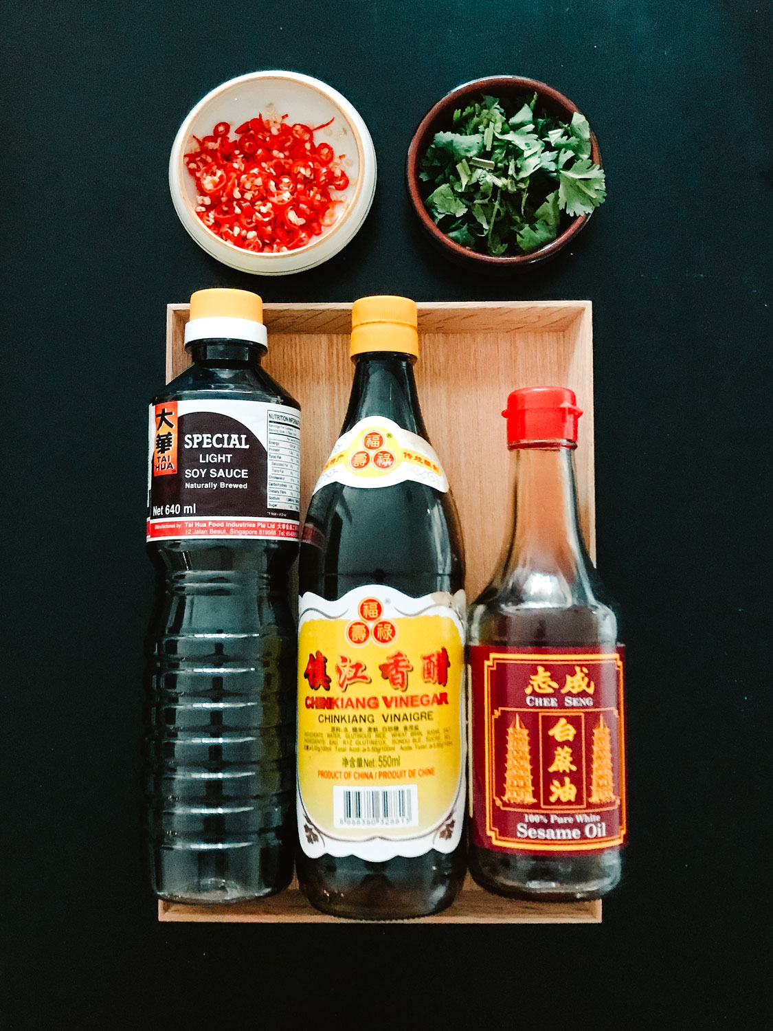 A bottle of sesame oil, soy sauce and black vinegar with small dishes of chili rings and coriander.
