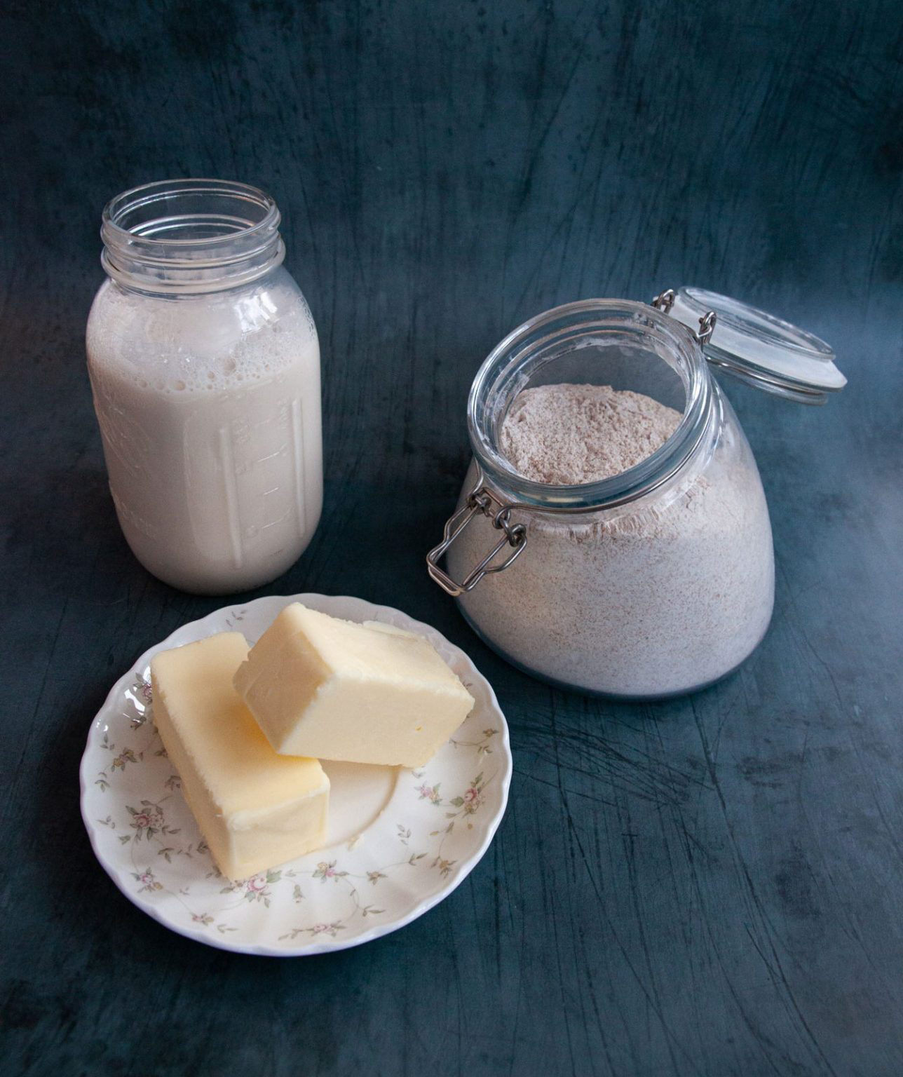 A small jar of milk, a plate of butter and a container of flour resting on a table.
