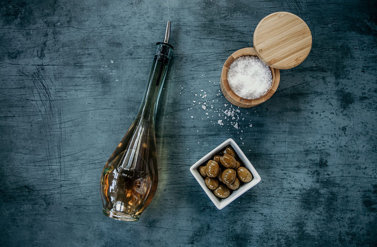 A sleek, skinny bottle of vinegar next to a dish of olives and a wooden container of kosher salt.