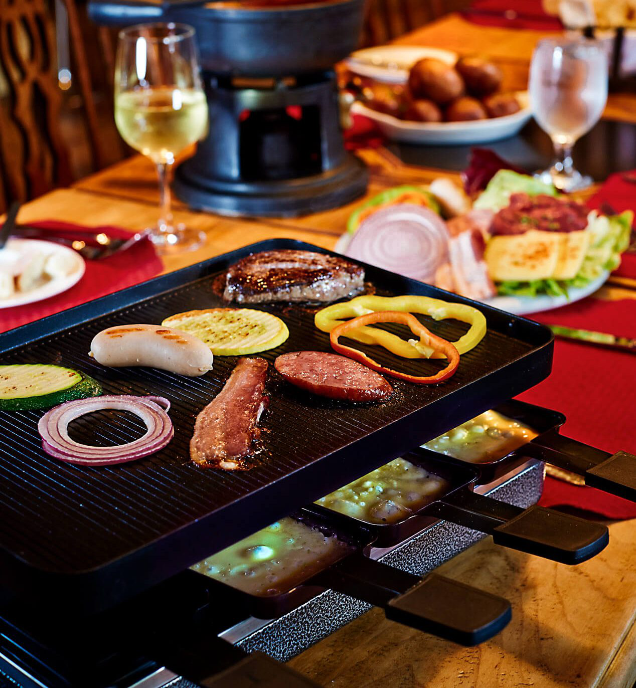 A raclette grill with a griddle on top cooking various meats and vegetables.