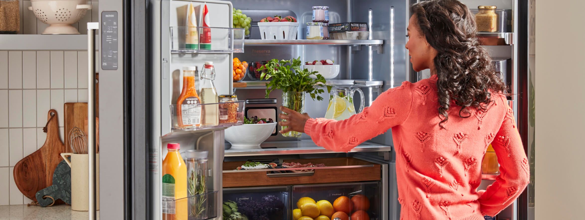 Woman reaching into KitchenAid® French Door Refrigerator to grab a jar holding fresh tender herbs.