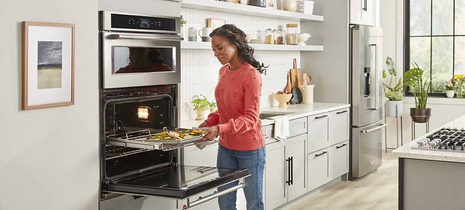 Woman removing a sheet pan from the oven in a bright kitchen with light gray cabinets.