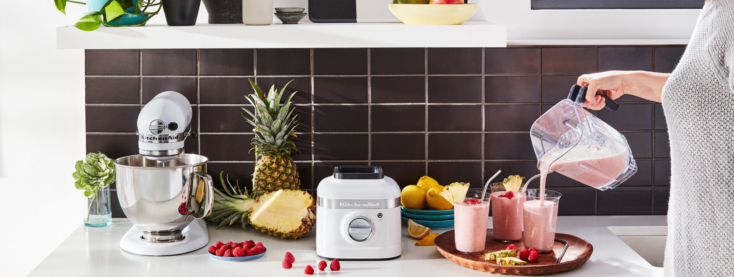  White countertop with KitchenAid® stand mixer and KitchenAid® blender surrounded by fruits - woman is pouring smoothies into three glasses.