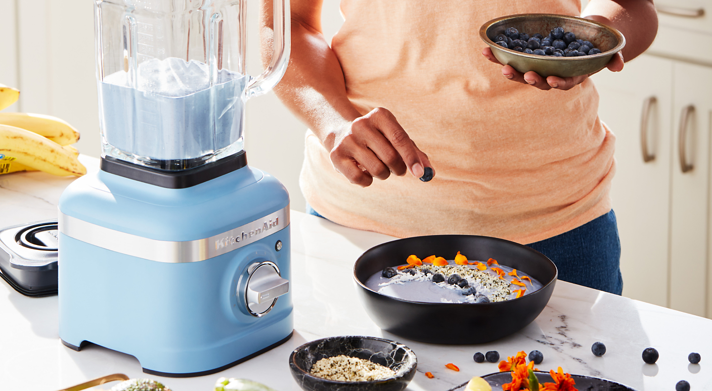 Person garnishing a smoothie bowl with blueberries next to a blue KitchenAid® blender