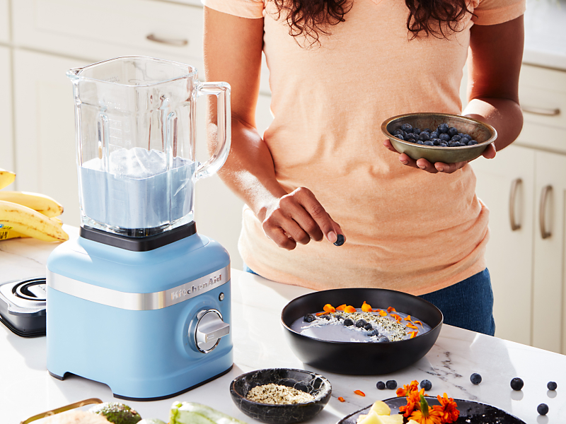 Person garnishing a smoothie bowl with blueberries next to a blue KitchenAid® blender
