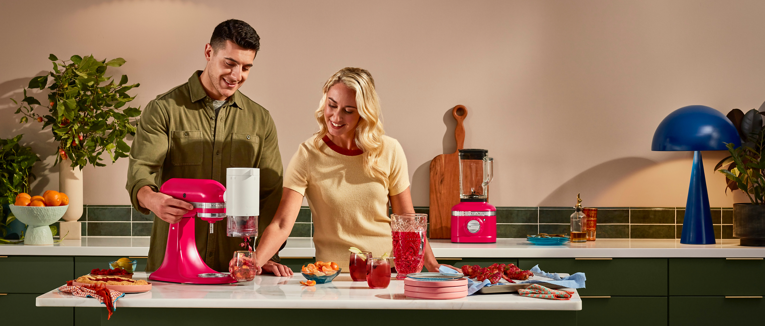 A man and woman in a stylish kitchen using a Hibiscus Stand Mixer with the Shaved Ice Attachment to make a colorful beverage, and a Hibiscus Blender on a counter behind them. 