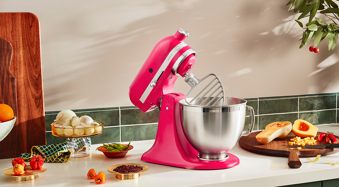 A Hibiscus Stand Mixer on the countertop surrounded with different fresh ingredients in stylish, modern bowls and on wood cutting boards. 