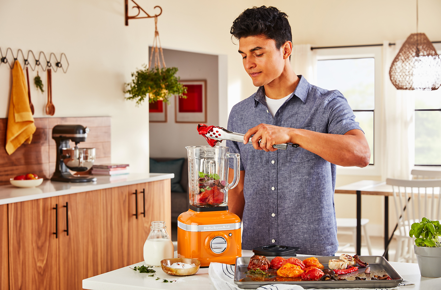 A man adding roasted vegetables to a KitchenAid® blender in a modern kitchen.