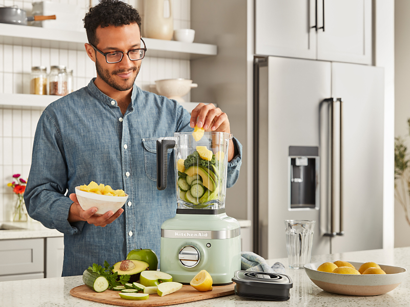 A man putting fresh vegetables into a KitchenAid® blender in a modern kitchen.