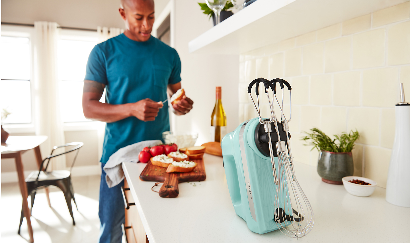 A man in a modern kitchen spreading goat cheese on a crostini next to a KitchenAid® hand mixer. 