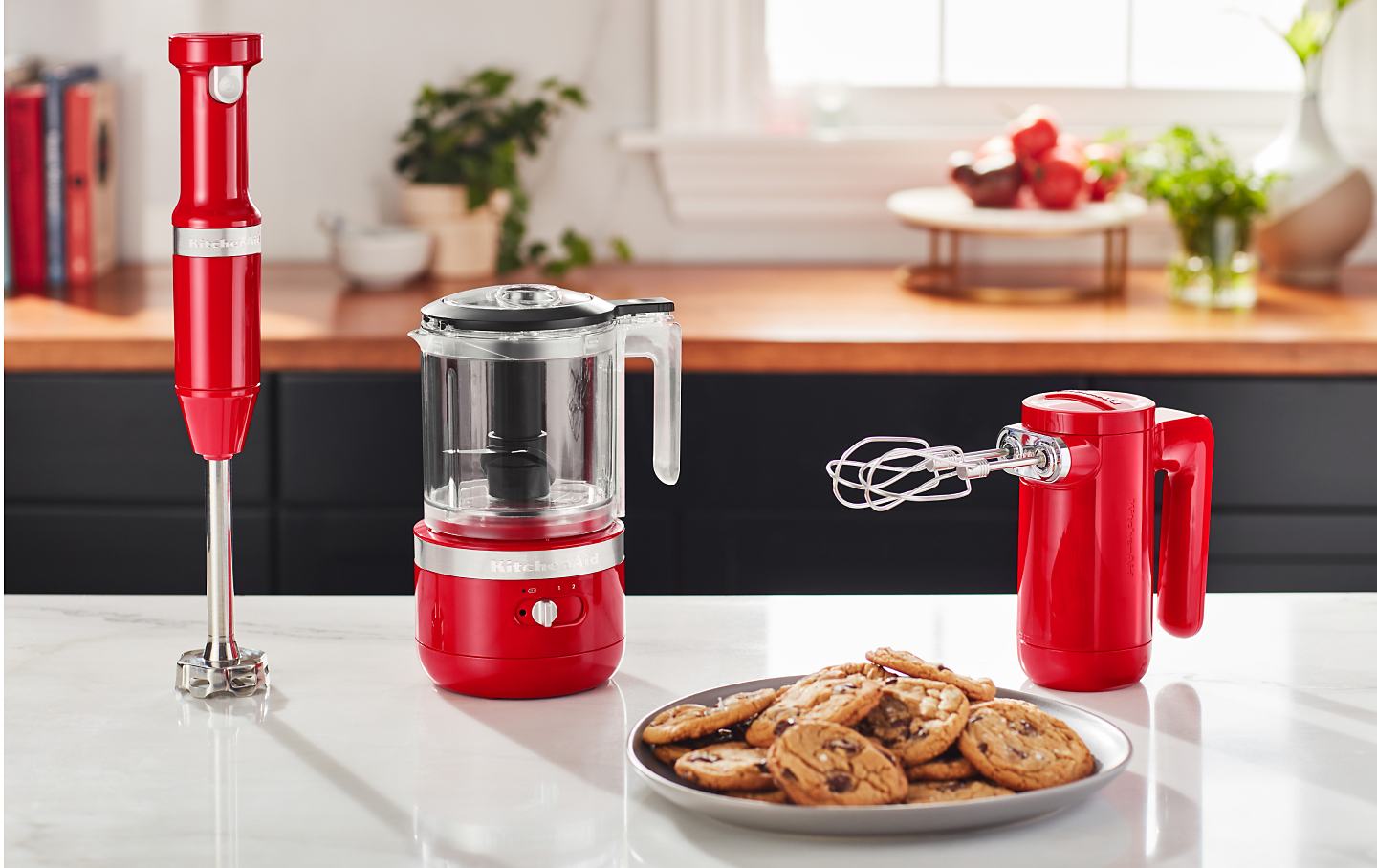 Cordless KitchenAid® hand mixer, hand blender and food chopper next to a plate of freshly baked chocolate chip cookies.