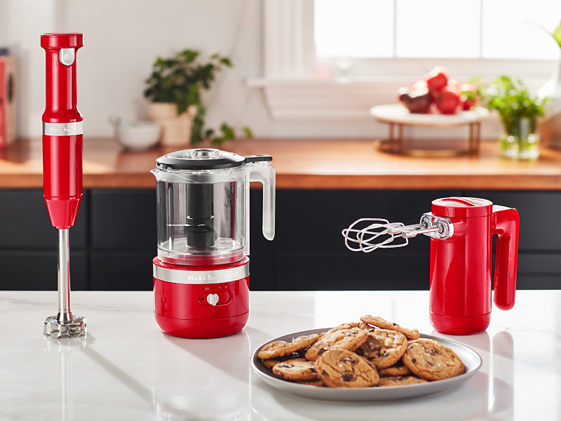 tbaCordless KitchenAid® hand mixer, hand blender and food chopper next to a plate of freshly baked chocolate chip cookies.
