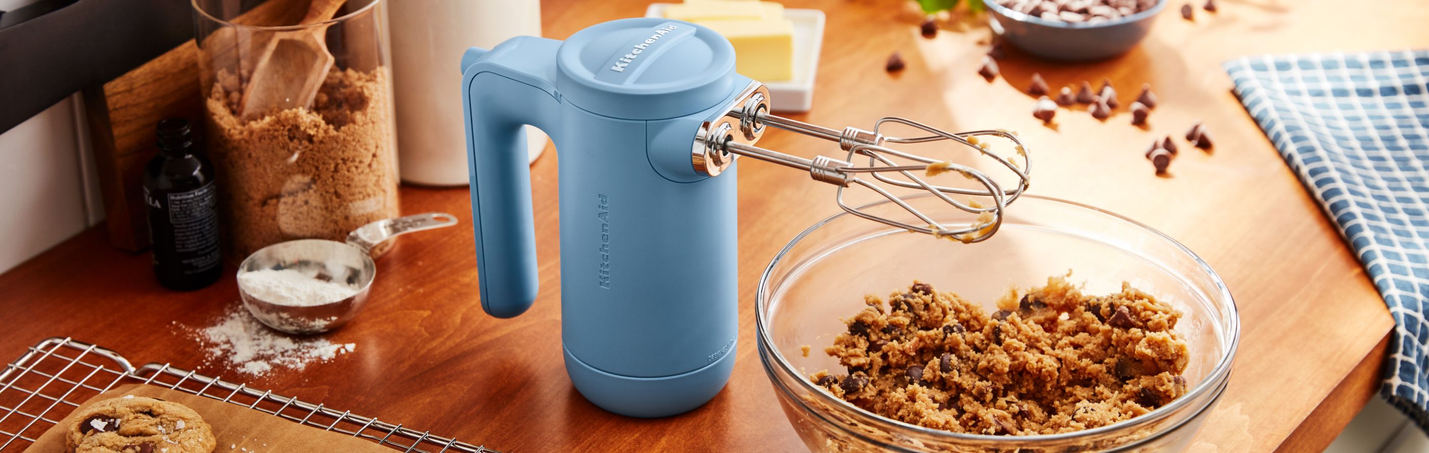 Cordless KitchenAid® hand mixer next to cookie dough and fresh baked chocolate chip cookies.
